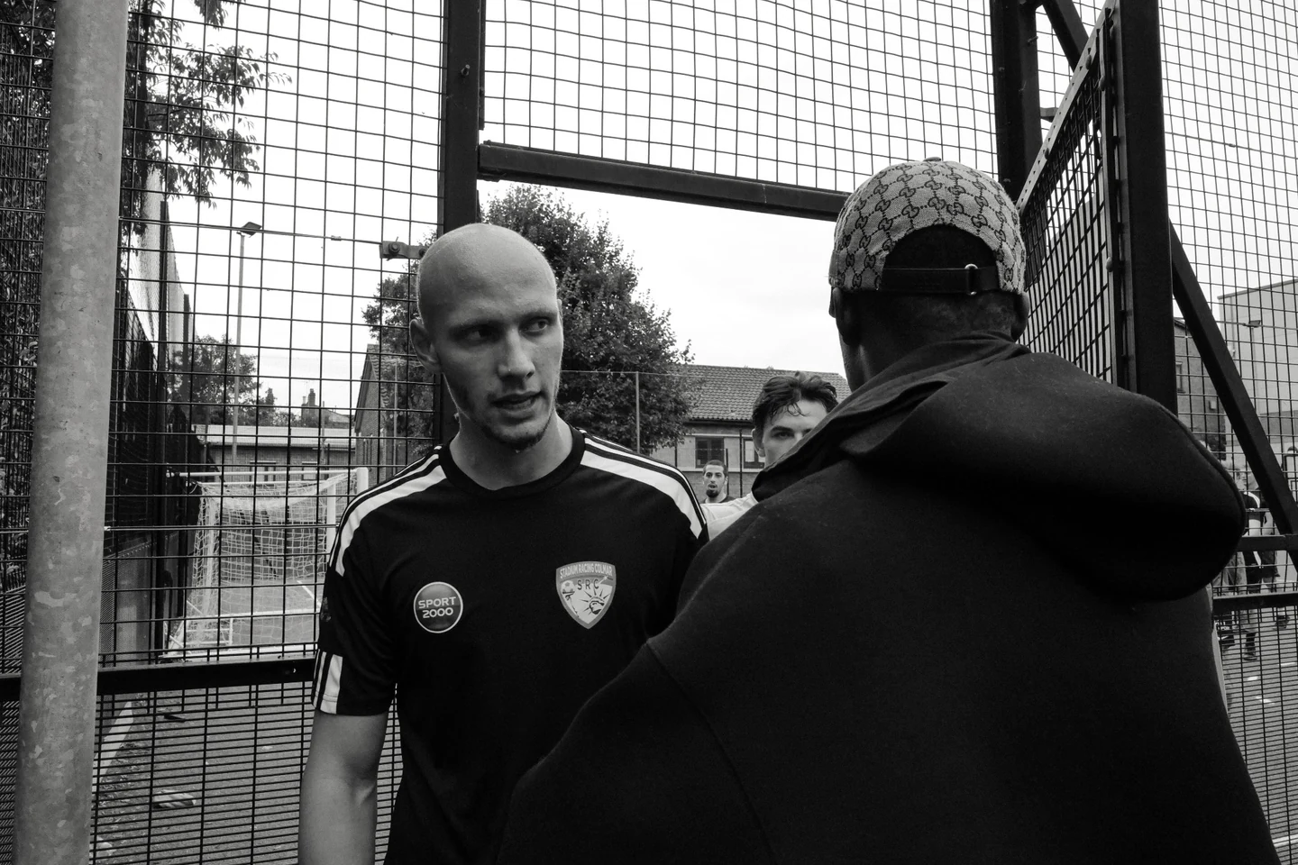 Two footballers talking beside the cage of a small-sided pitch.